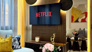 Un salon moderne doté d’un grand téléviseur à écran plat affichant le logo « NETFLIX », d’un canapé agrémenté de coussins à motifs, d’une table basse sur laquelle est posé un vase de fleurs et d’une sculpture décorative.