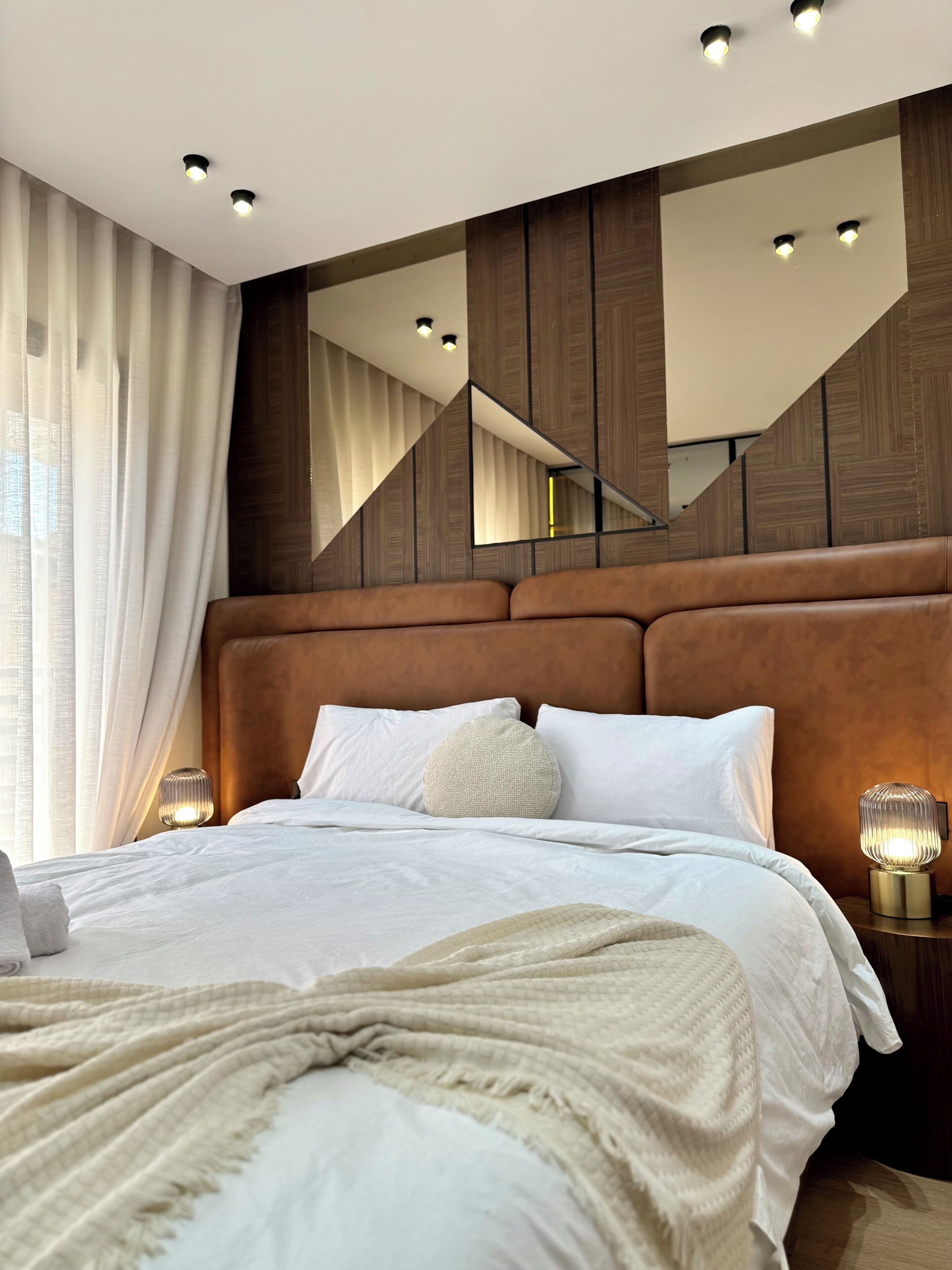 Une chambre moderne avec un grand lit, une tête de lit dotée d’un éclairage intégré, un miroir et une table de chevet avec une lampe.