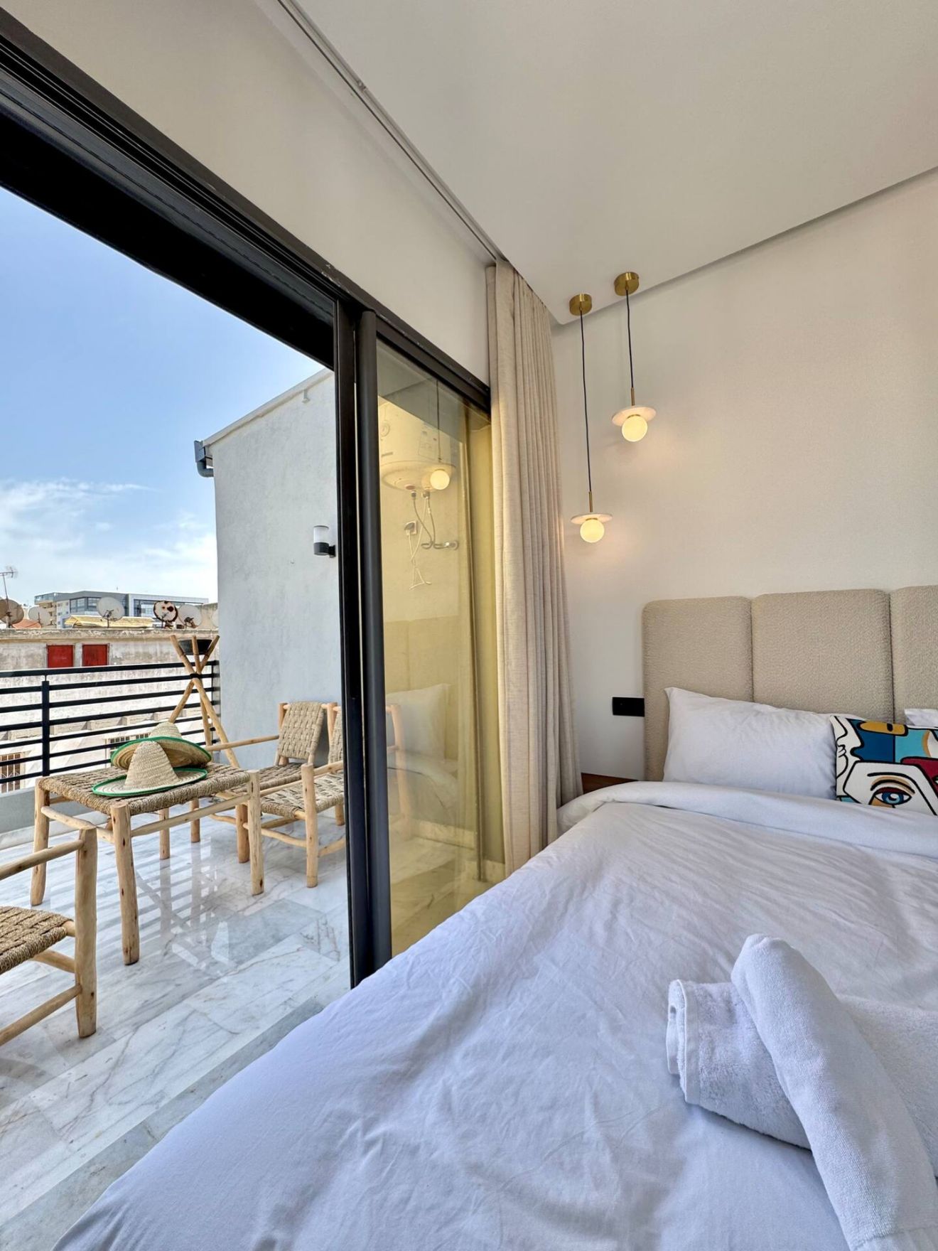 A modern hotel room with a large bed, a balcony with wicker chairs, and a view of the city.