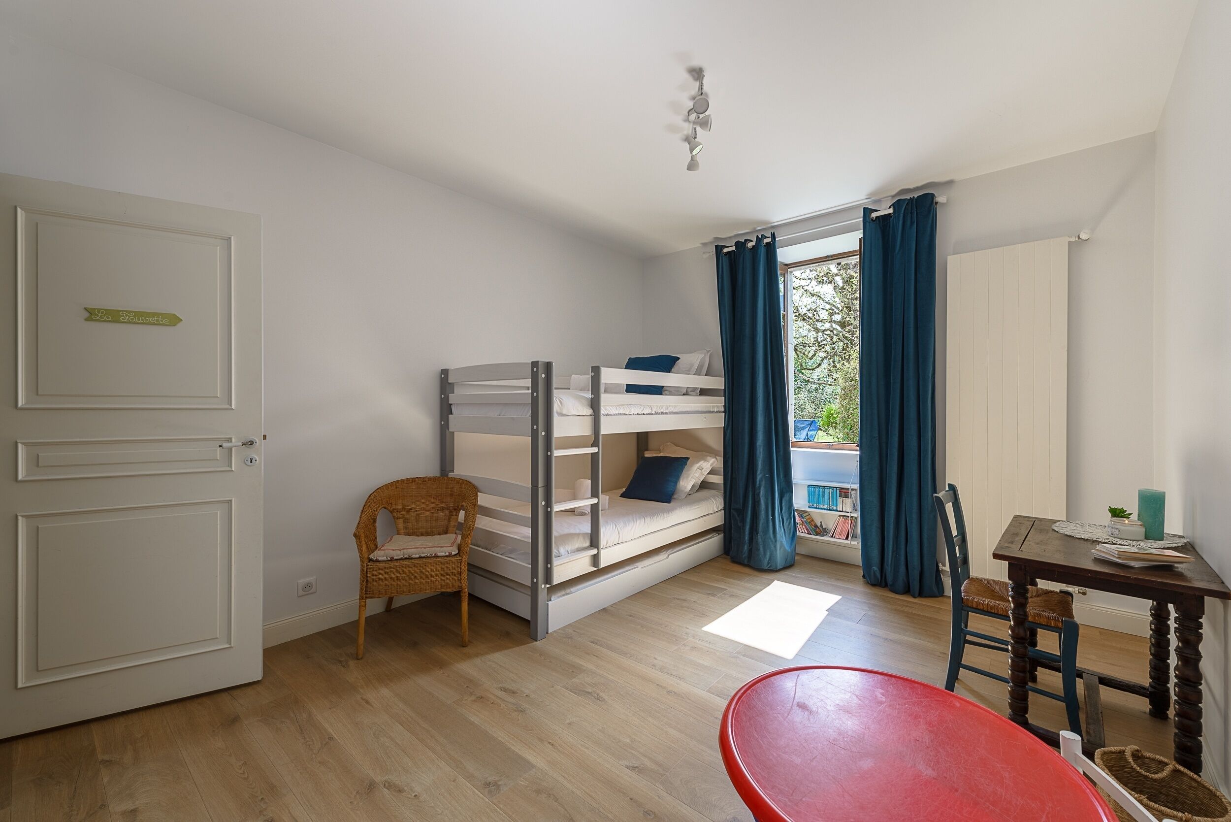 A room with a bunk bed, a wicker chair, a red table, and a view of greenery outside.