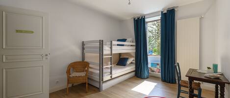 A room with a bunk bed, a wicker chair, a red table, and a view of greenery outside.