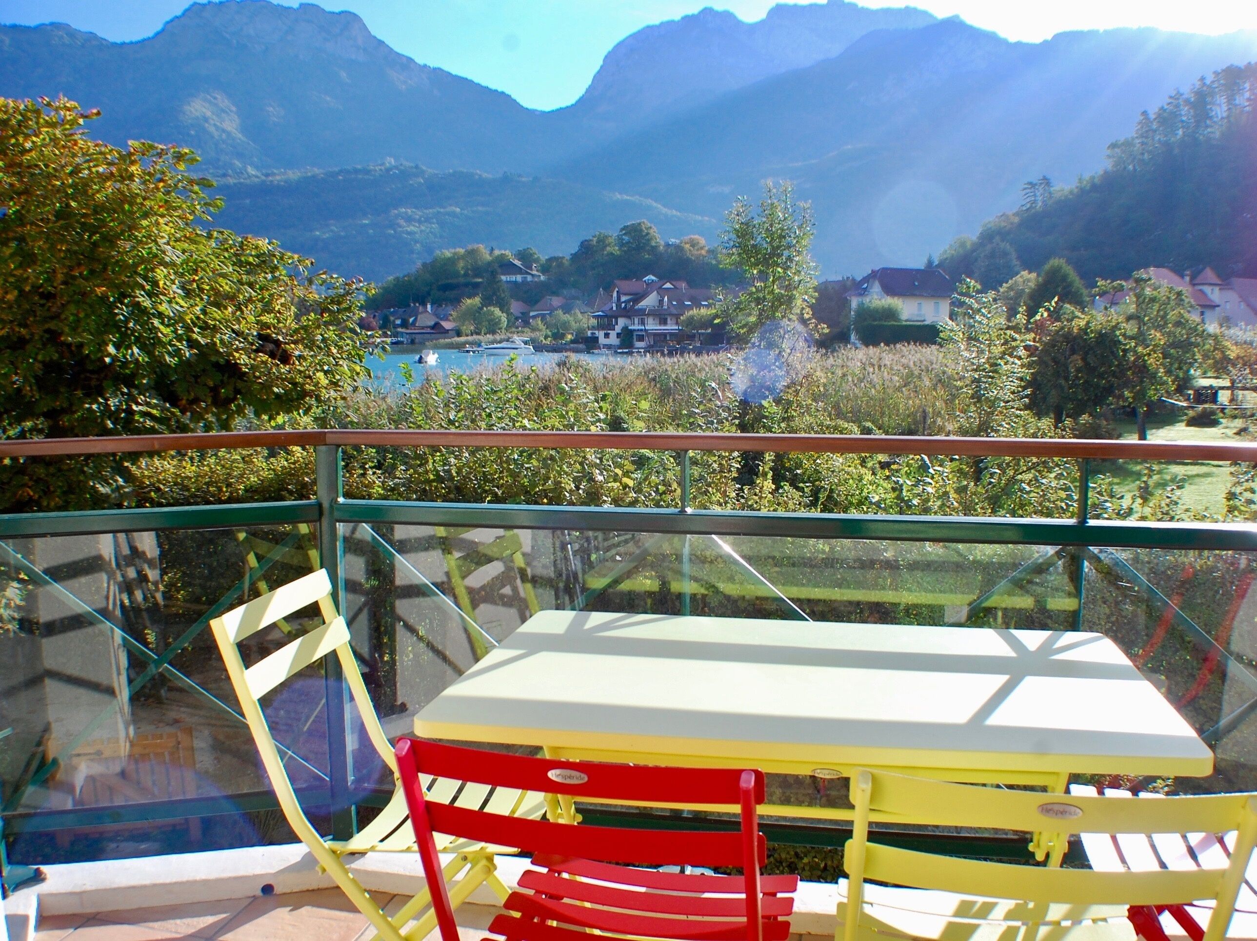 A balcony with a table and chairs, overlooking a mountainous landscape with a lake and buildings.
