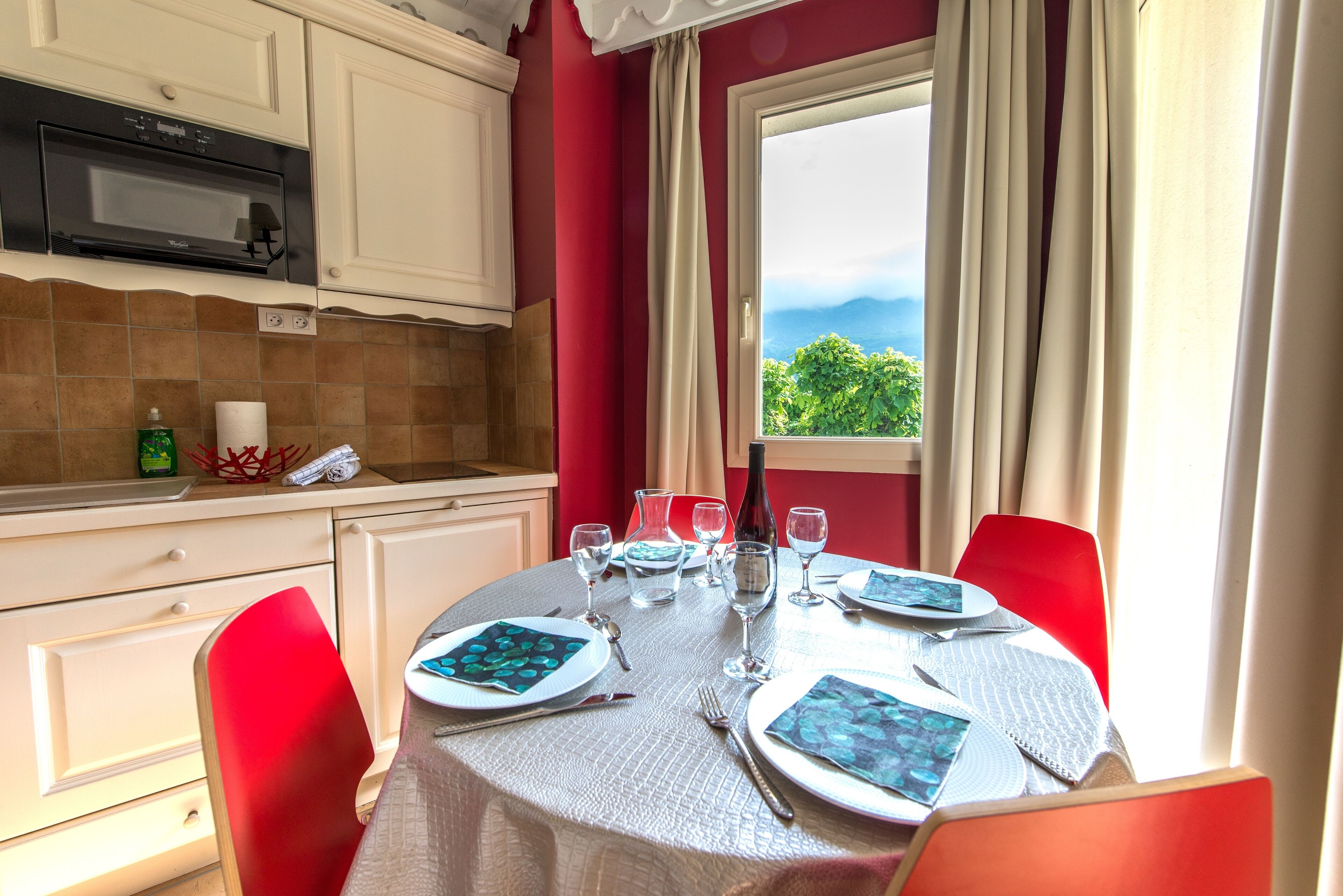 Une table à manger dressée avec des assiettes, des verres et des couverts, et une fenêtre donnant sur un paysage verdoyant et montagneux.