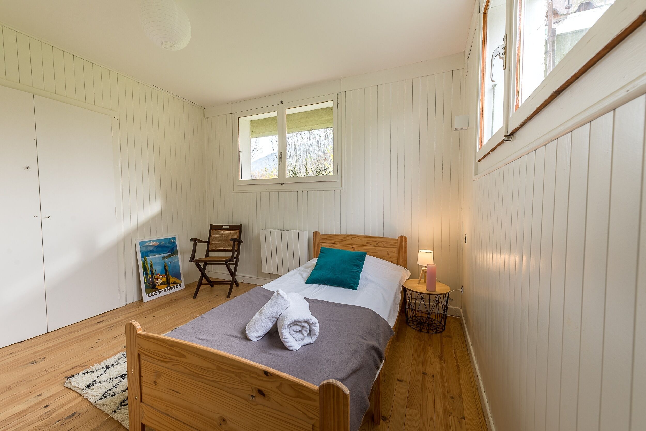Een slaapkamer met een houten bed, een raam, een stoel, een poster en een klein tafeltje met een lamp.