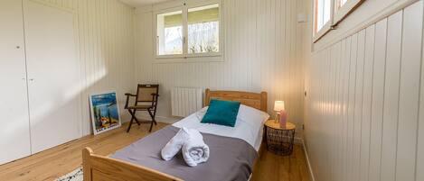 Een slaapkamer met een houten bed, een raam, een stoel, een poster en een klein tafeltje met een lamp.