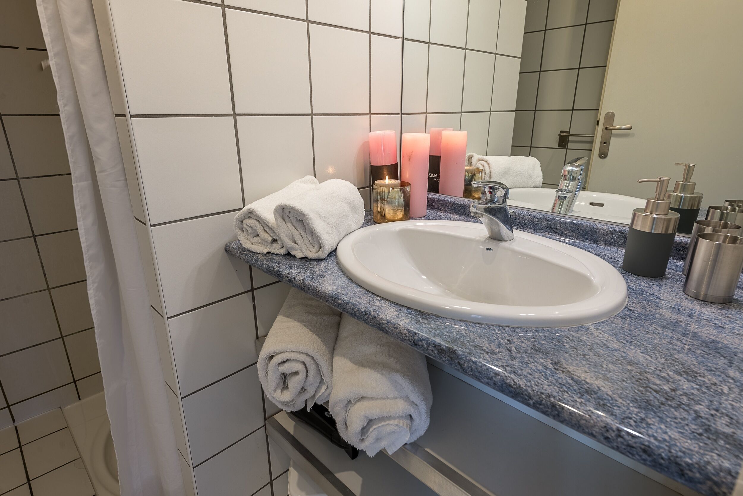 A bathroom with a white sink, marble countertop, and a mirror. There are three candles, a towel rack with folded towels, and a shower curtain.
