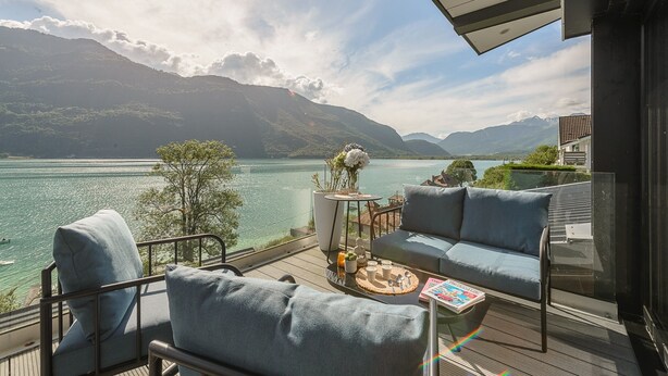 A balcony with a view of a lake and mountains, furnished with a sofa and chairs, and a small table with a vase and a magazine.