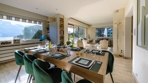 Un comedor moderno con una mesa de madera dispuesta para seis personas, rodeada de sillas tapizadas en verde. Se ve una estantería y un comedor con vistas a las montañas al fondo.