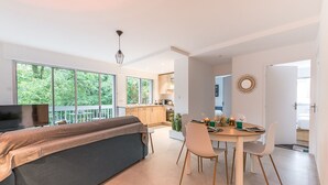 A modern kitchen with large windows, a dining area with a round table, and a sofa with a blanket.