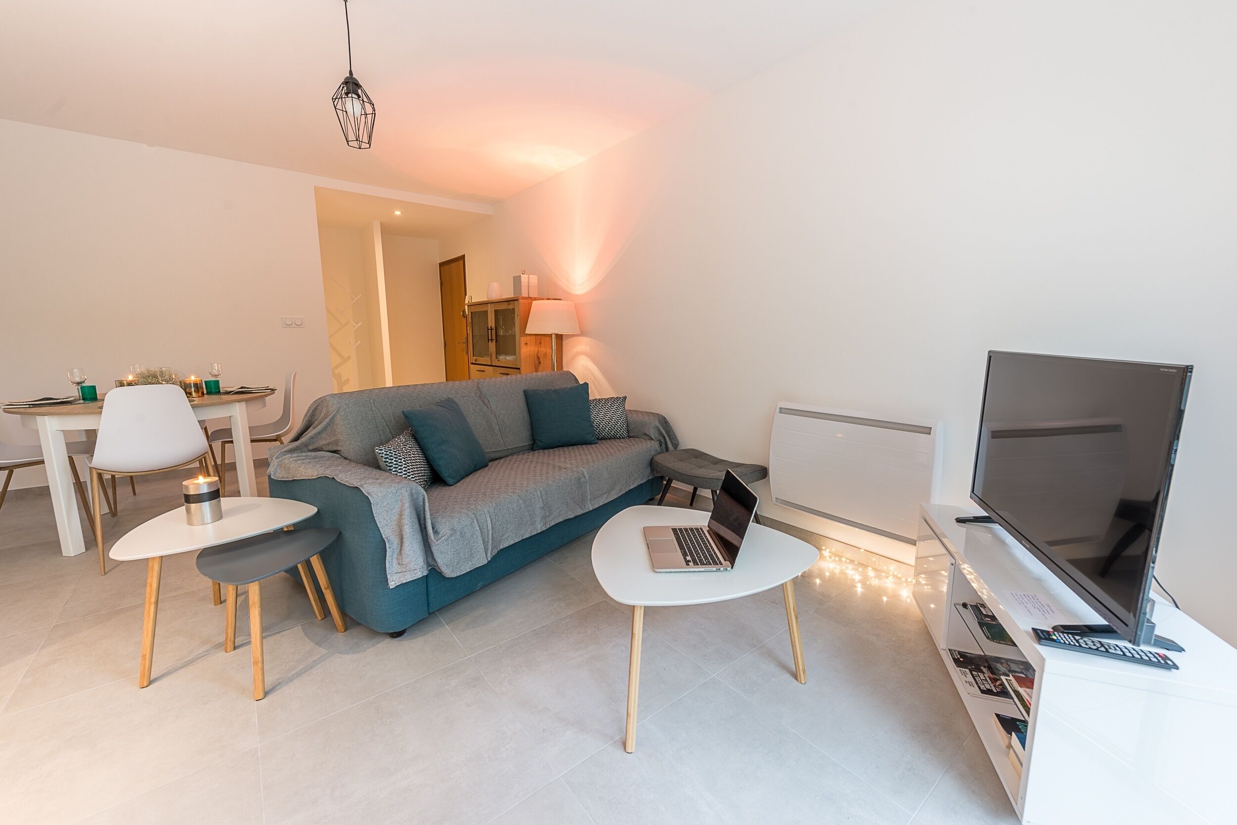 En moderne stue med en grå sofa, et hvidt sofabord, et fjernsyn og et spiseområde med et hvidt bord og stole.