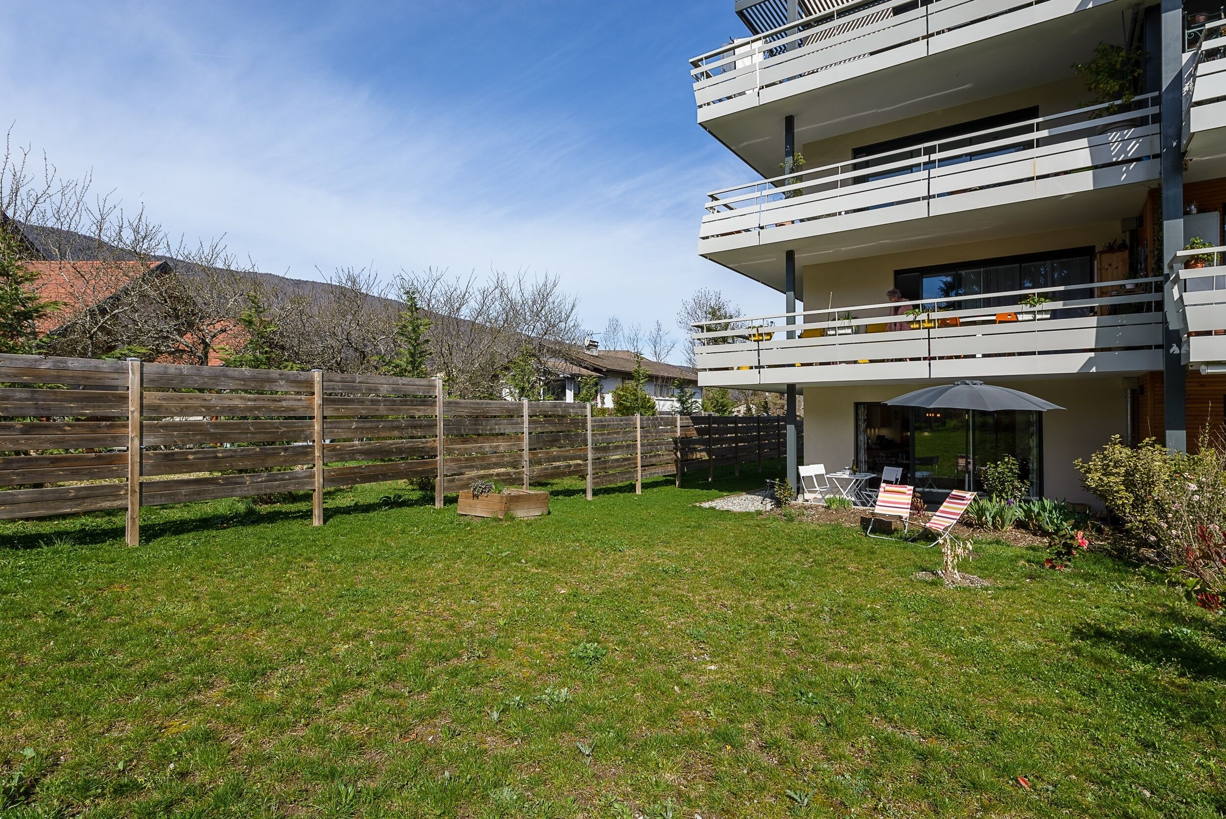 Un immeuble moderne à plusieurs étages, doté d’un balcon, d’une clôture en bois et d’une cour verdoyante aménagée avec du mobilier d’extérieur.