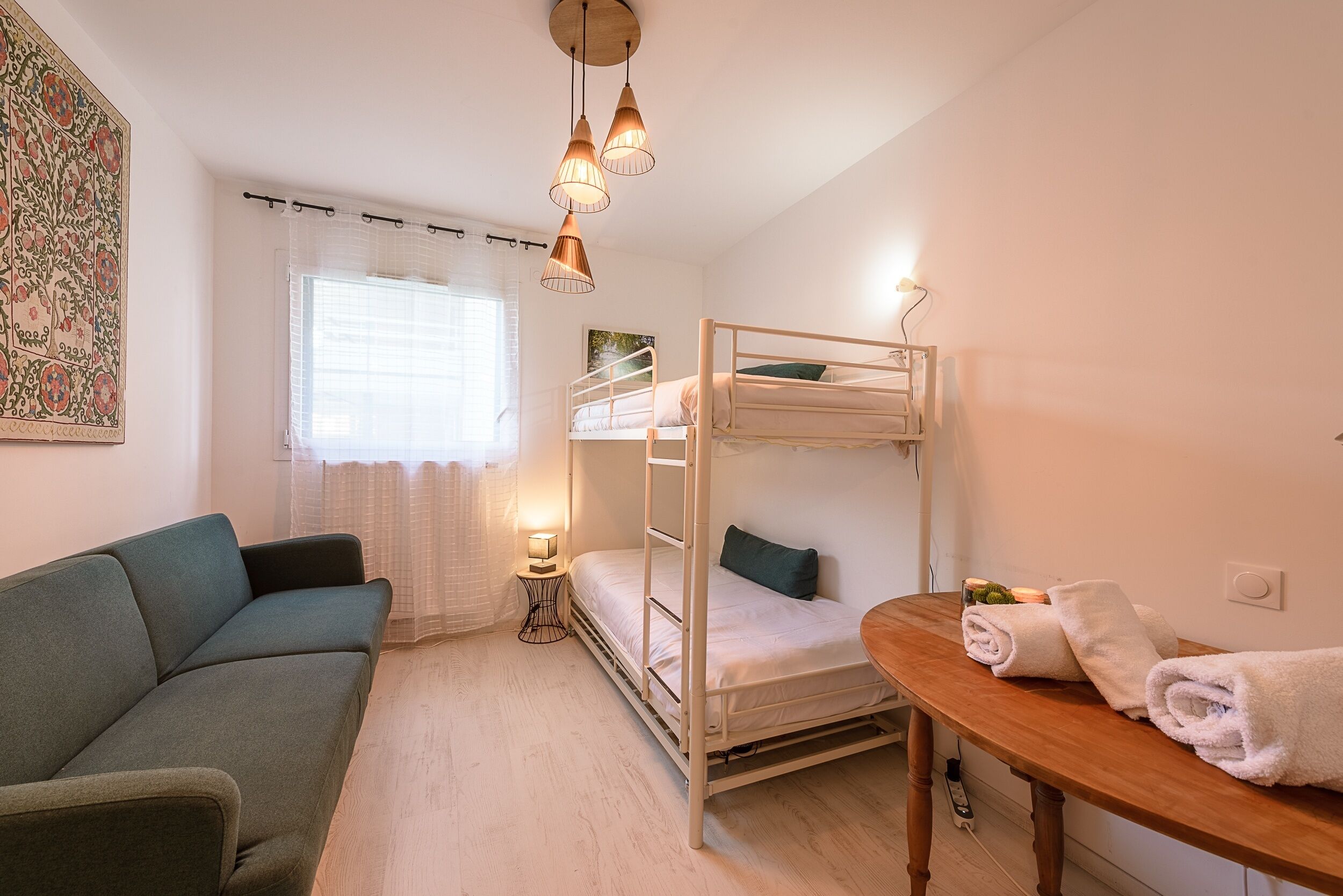 Una habitación pequeña y luminosa con una cama litera, un sofá, una mesa con toallas y una ventana con cortinas.