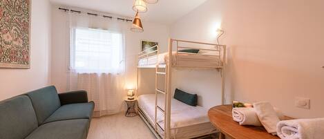 Una habitación pequeña y luminosa con una cama litera, un sofá, una mesa con toallas y una ventana con cortinas.