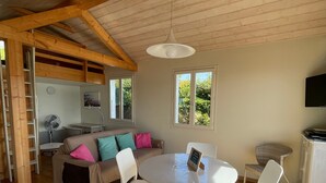 Una sala de estar acogedora con techo de madera, una mesa redonda blanca, sillas blancas, un sofá con cojines de colores, un televisor y vistas a zonas verdes.