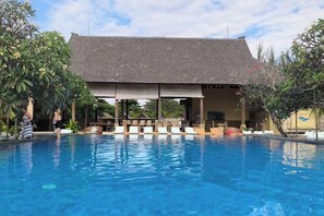 Une piscine avec des chaises longues, un espace de détente couvert et un bâtiment doté d’un toit en tuiles.