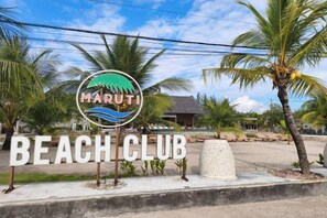 A beach club sign with a logo, palm trees, and a building in the background. - Maruti Beach Villas (Bali)