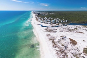 An aerial view of a coastal area with a beach, a cluster of buildings, and a clear view of the ocean.