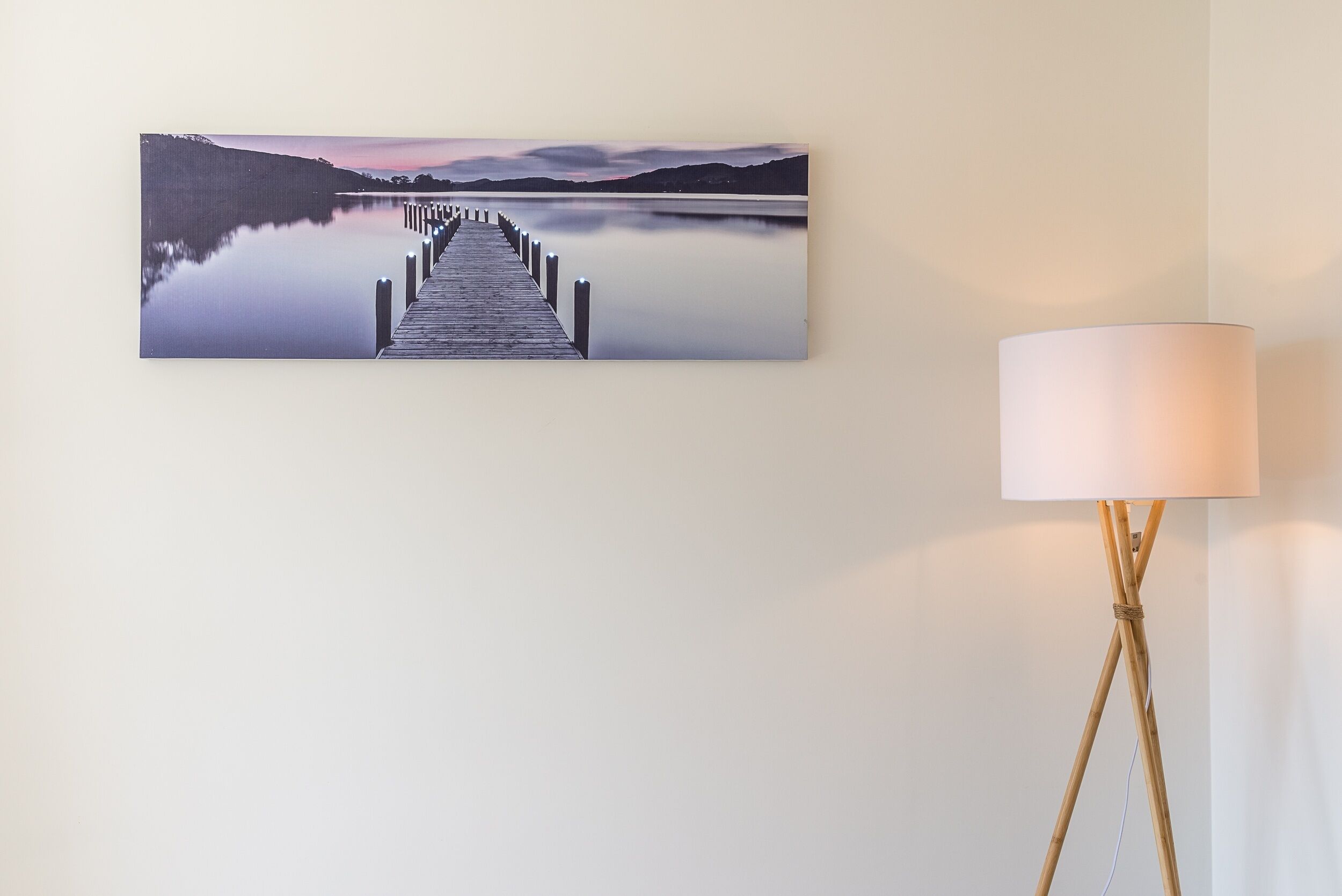Une peinture représentant un paysage, encadrée, et montrant une jetée s’avançant dans un lac paisible, avec des montagnes en arrière-plan.