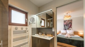 A modern bathroom with a shower, a toilet, and a sink. There is a mirror above the sink and a window above the shower. The bathroom has a wooden door and a towel rack.