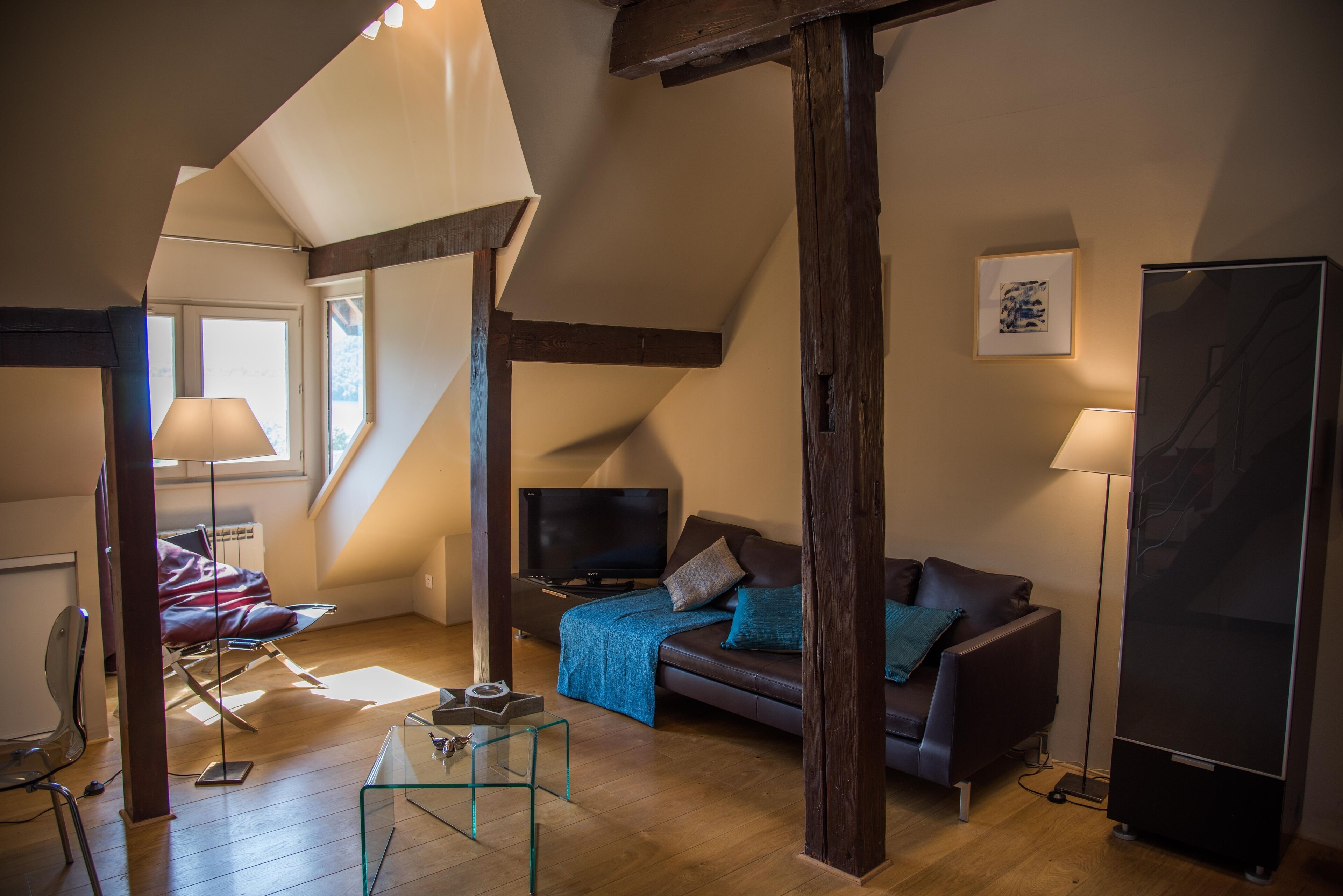 A loft-style living room with a high ceiling, exposed wooden beams, a sofa, a TV, and a glass coffee table.
