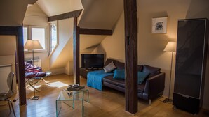 Een loft-achtige woonkamer met een hoge plafond, zichtbare houten balken, een bank, een televisie en een glazen salontafel.