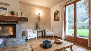 Ein gemütliches Wohnzimmer mit Kamin, einem Esstisch mit Gebäck, einer bequemen Couch und Blick durch eine Glastür.