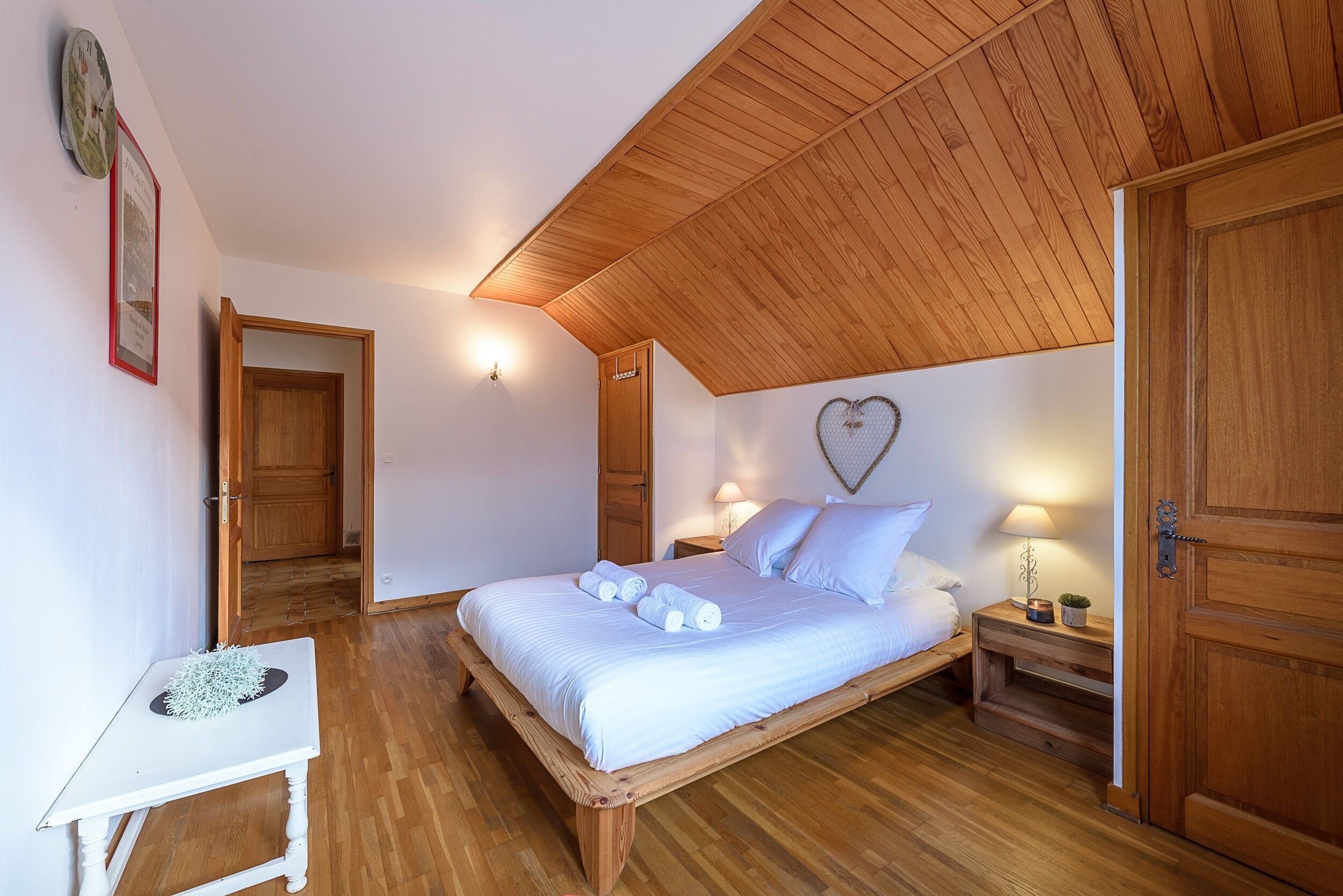 A bedroom with a wooden ceiling, a bed with white linens, a bedside table with a lamp, and a heart-shaped decoration on the wall.