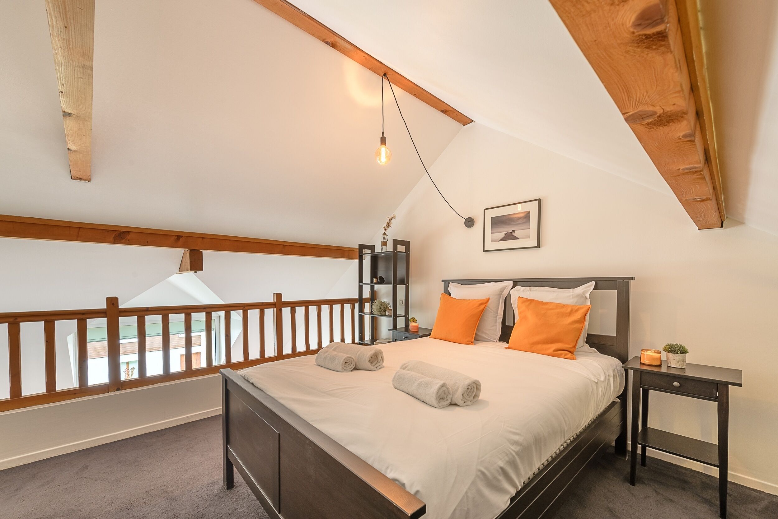 A bedroom with a wooden ceiling, a bed with white and orange bedding, a nightstand with a plant, and a view of the outdoors.
