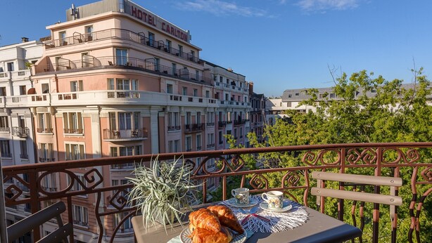 A balcony with a table set for breakfast, including croissants and coffee cups, overlooking a cityscape with multi-story buildings.