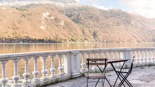 A lakeside view with a wooden table and chairs on a balcony with a white balustrade.