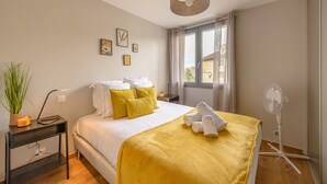 A hotel room with a bed, a bedside table with a lamp, a fan, and a window with curtains.