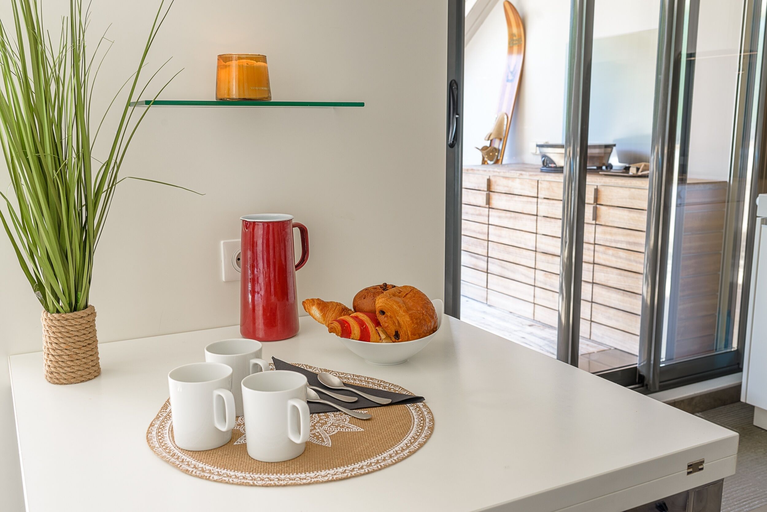 Une table dressée pour le petit-déjeuner, avec une nappe blanche, une tasse rouge, un bol de croissants et une plante en pot.
