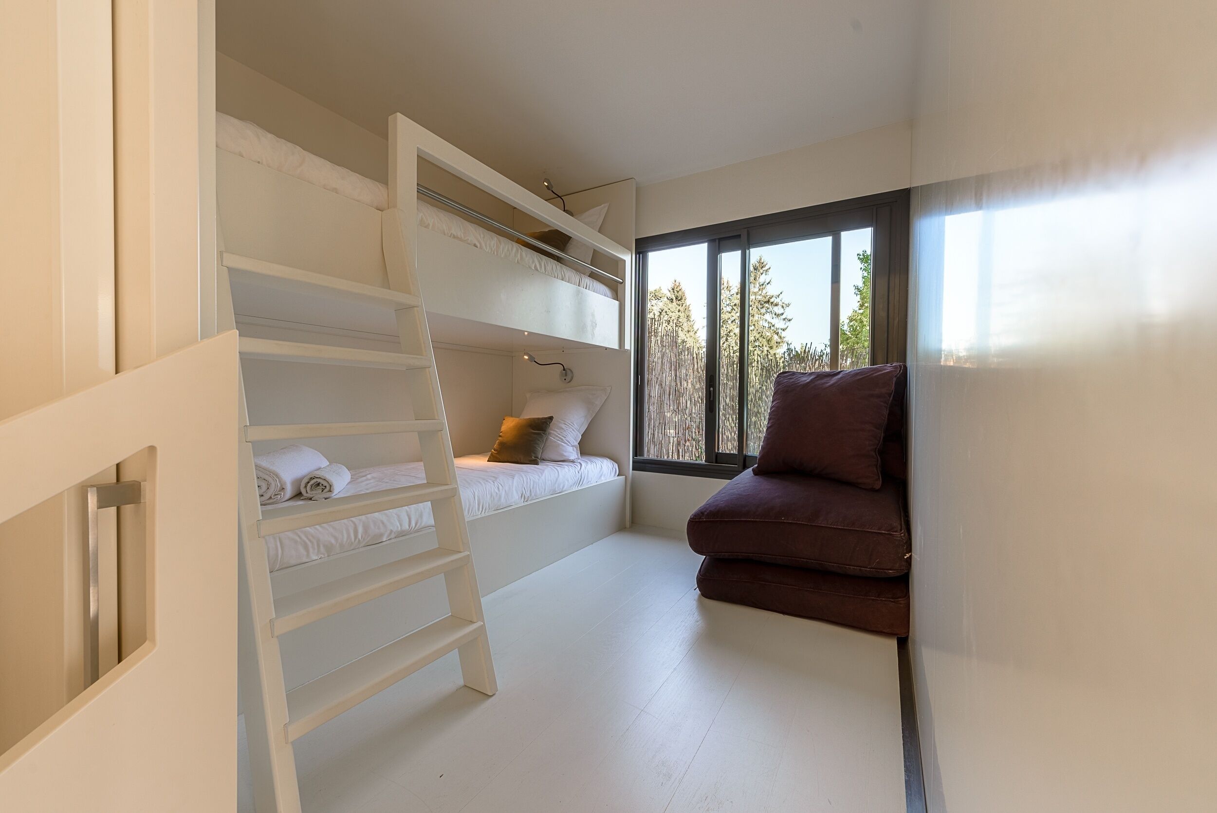 Habitación con literas, una escalera, un ventanal con vistas a árboles y una silla marrón.