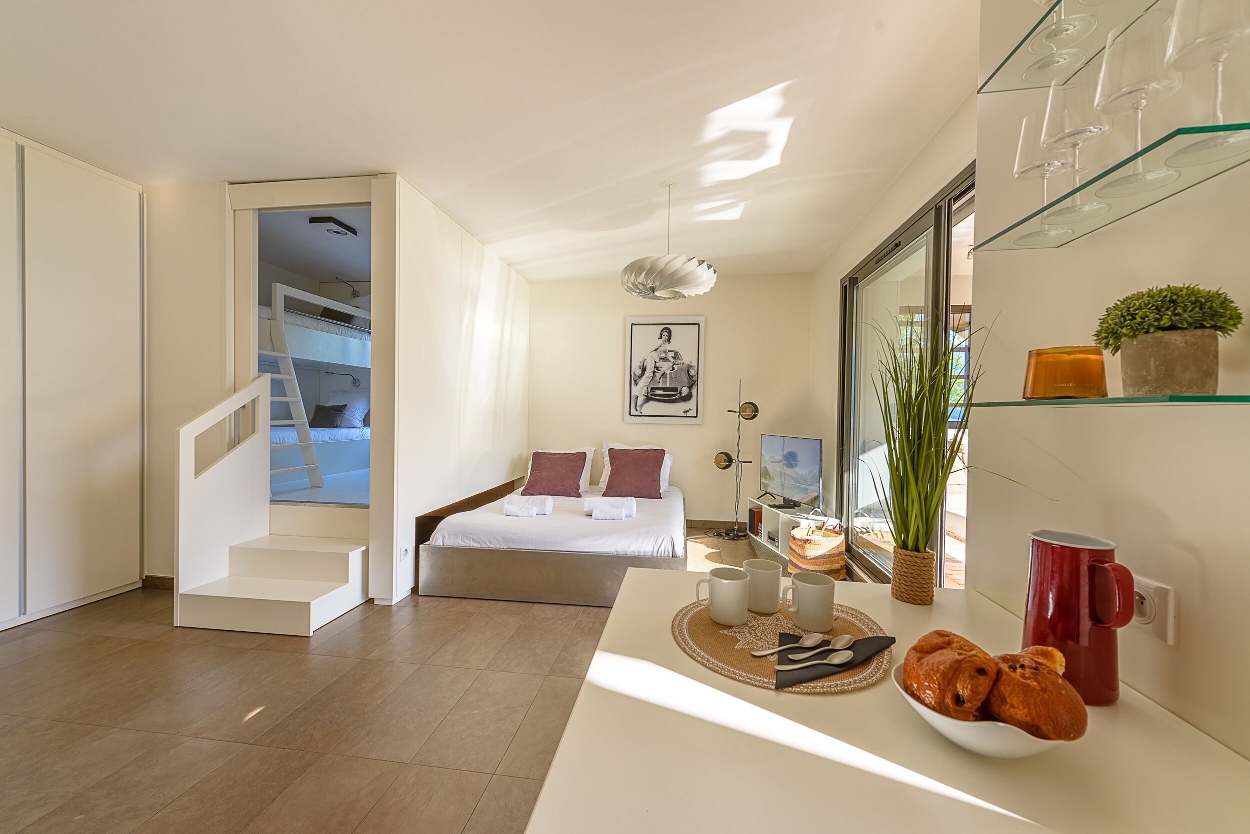 Ett modernt hotellrum med en säng, ett litet bord med ett fat fullt av bakverk, en röd mugg och en växt på en hylla.