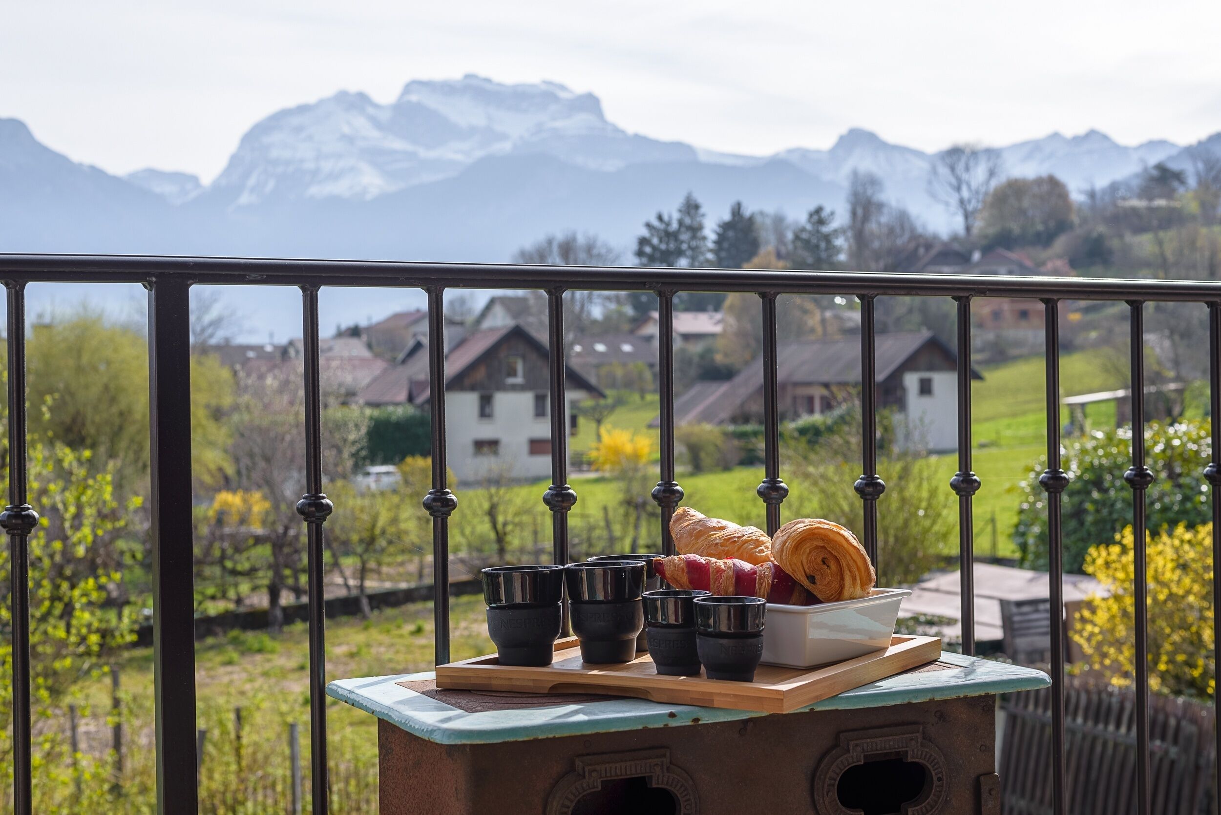 Un balcón con vistas a montañas, un croissant y tazas negras sobre una tabla de madera.