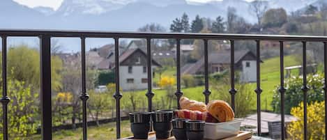 Un balcón con vistas a montañas, un croissant y tazas negras sobre una tabla de madera.