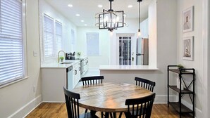 A dining area with a round wooden table and black chairs, a pendant light, and a black metal shelf with items on it. - Bright & Modern Prime Location (Jacksonville)