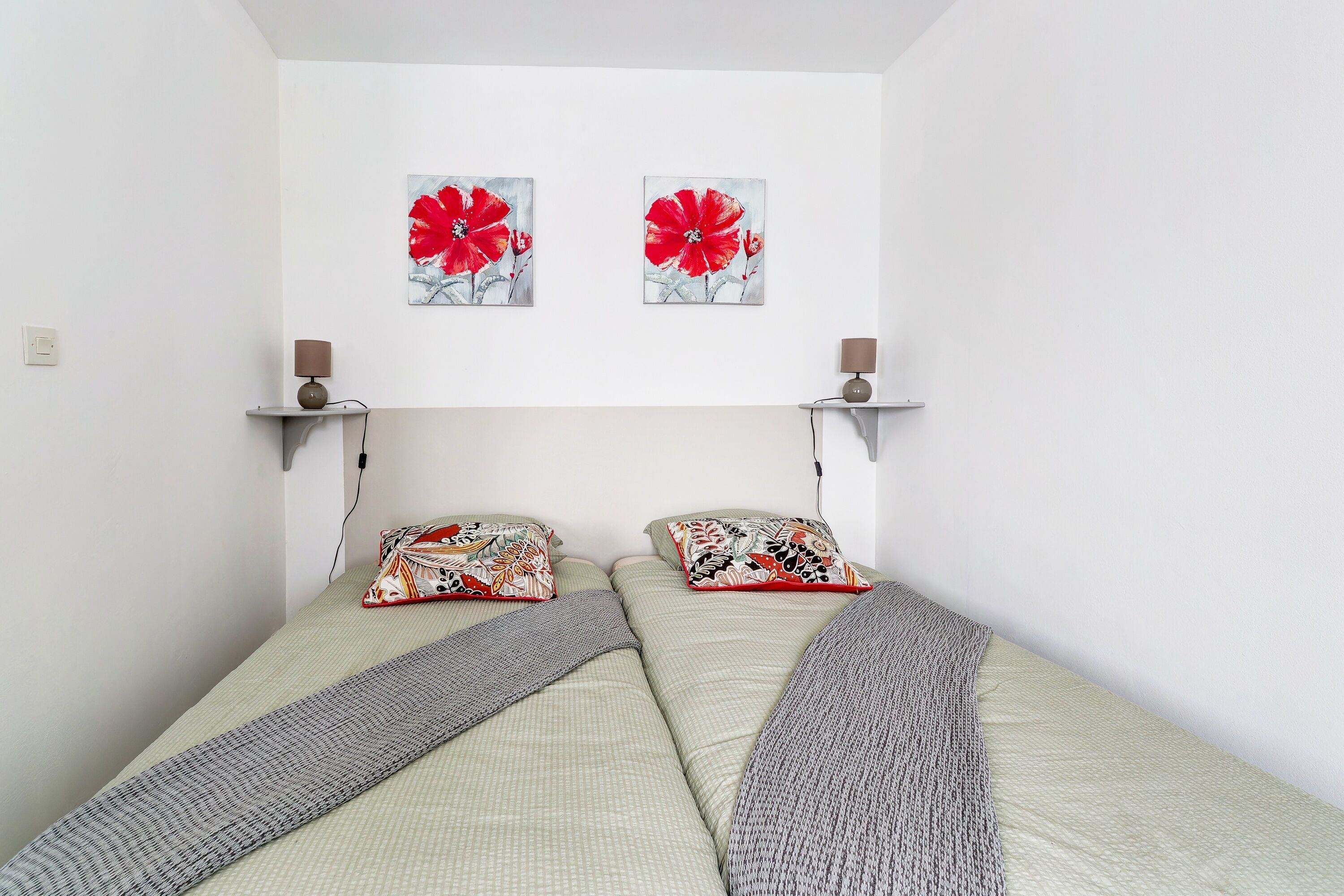 A bedroom with two beds, each with patterned pillows and a blanket. There are two wall-mounted lamps on either side of the beds.