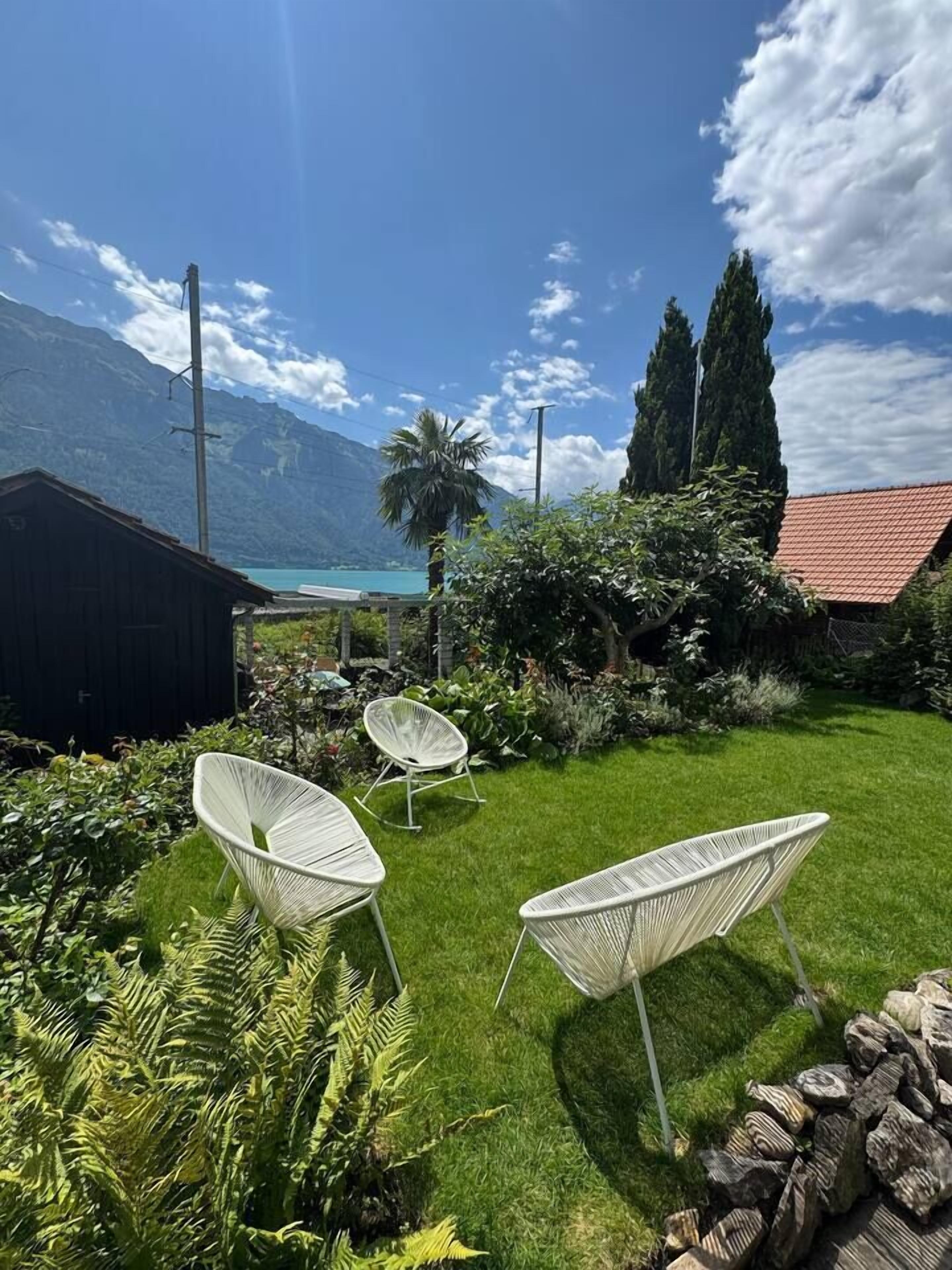 Un jardín con sillas blancas, un cobertizo de madera y vistas a una montaña y un lago.