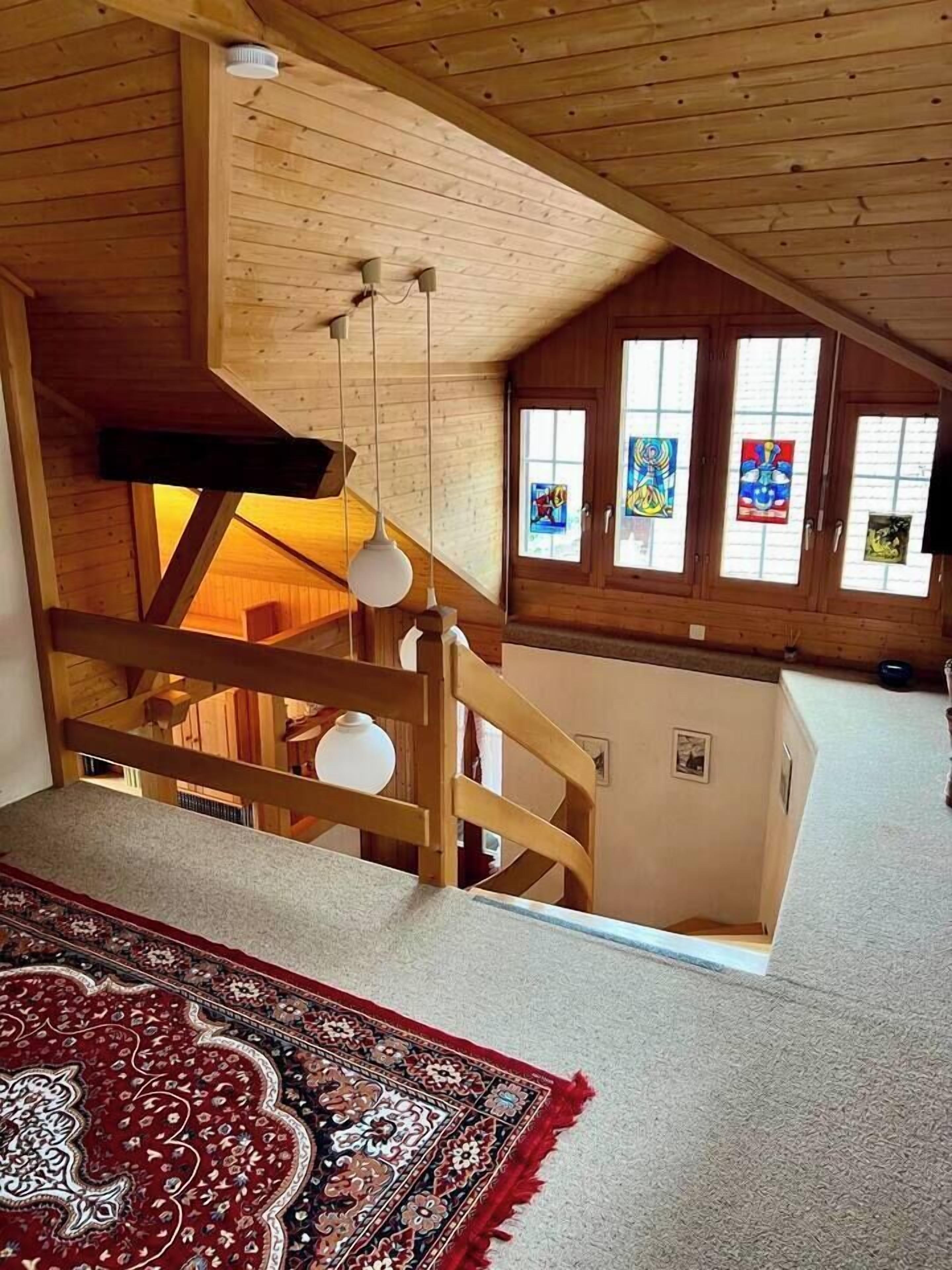 A wooden interior with a staircase, a red patterned rug, and a ceiling with hanging light fixtures.