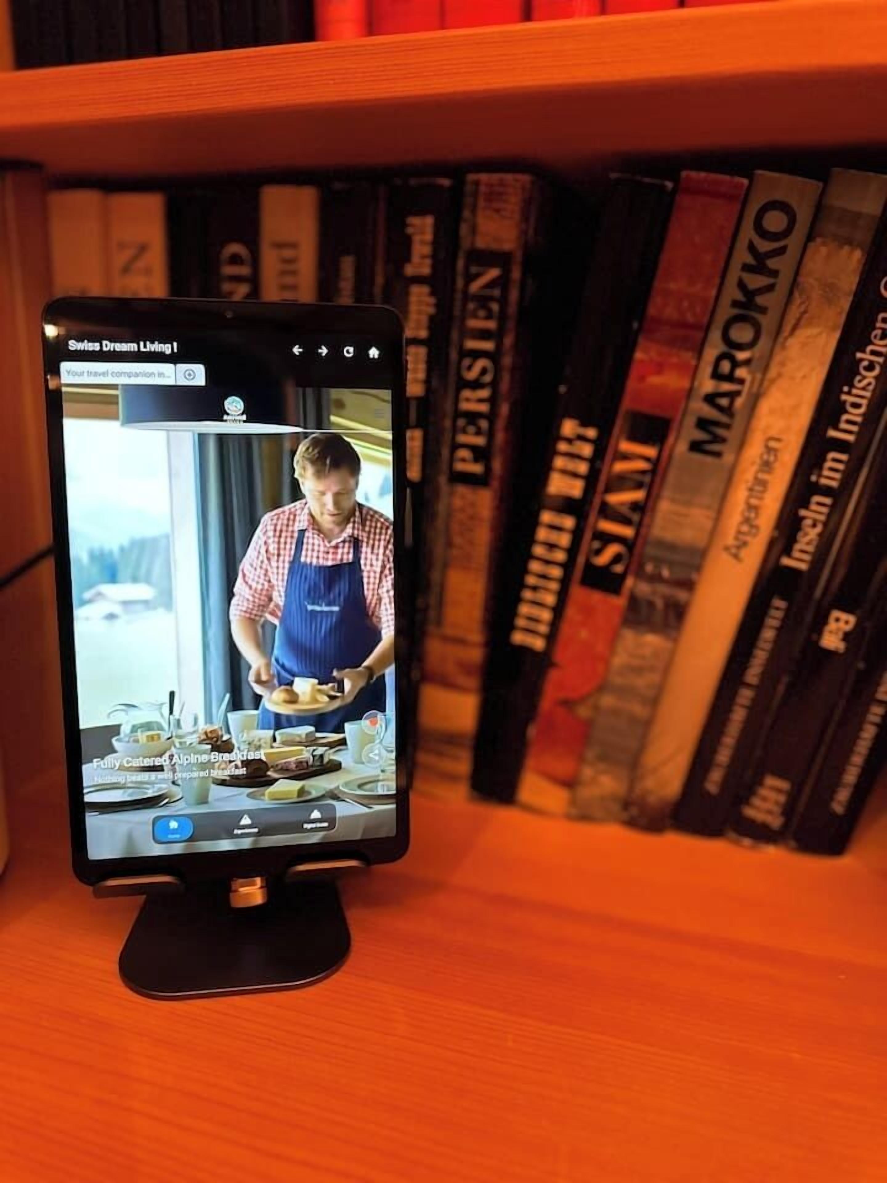 Una tableta muestra un video titulado 'Swiss Dream Living' con un chef preparando comida, sobre una superficie de madera con libros al fondo.