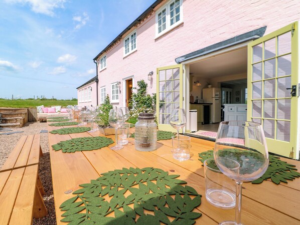 A wooden table set with wine glasses and a green leaf-patterned table runner, a rustic outdoor seating area, and a pink building with a kitchen visible through an open door.