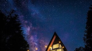 A triangular wooden cabin with large windows and a deck, set against a starry night sky.