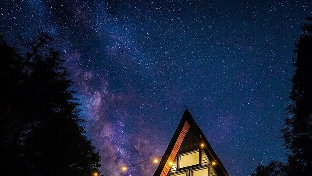 Una cabaña de madera triangular con ventanas amplias y una terraza, al pie de un cielo estrellado.