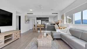 Een moderne woonkamer met een grijze bank, een houten salontafel en een televisie. Op de achtergrond is een keuken met grijze kastjes en een eethoek met houten stoelen zichtbaar.