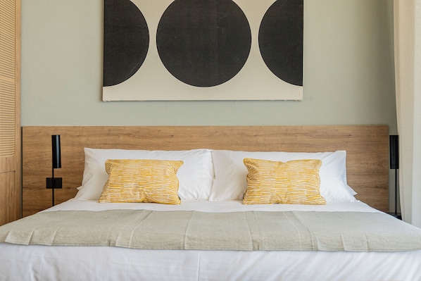 A neatly made bed with white bedding and two striped pillows, a wooden headboard, and a large abstract painting on the wall. - CasaChica Hotel (José Ignacio)