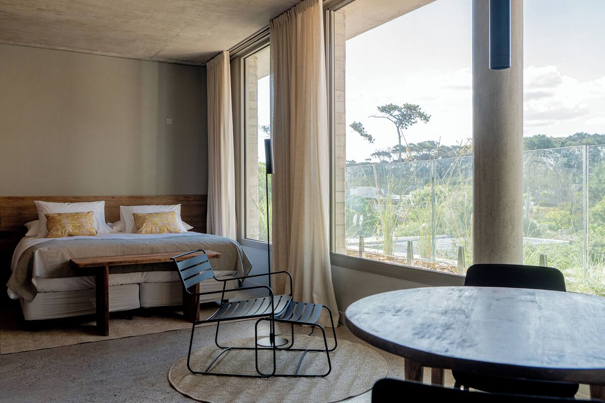 A hotel room with a bed, a wooden bench, a round table, and a view of greenery outside.