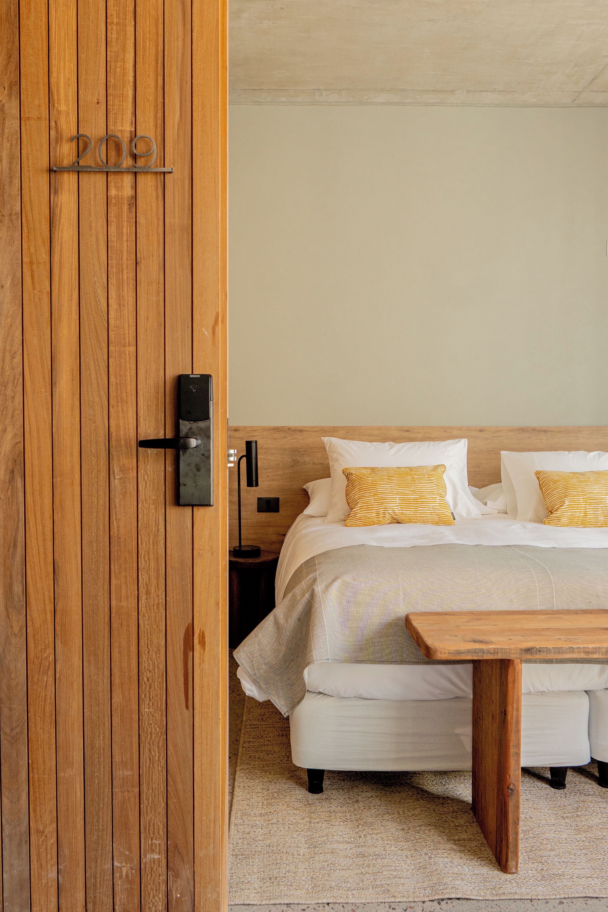 Una puerta de madera con el número 209, una cama con ropa de cama blanca y almohadas a rayas, una mesita de noche y una lámpara.