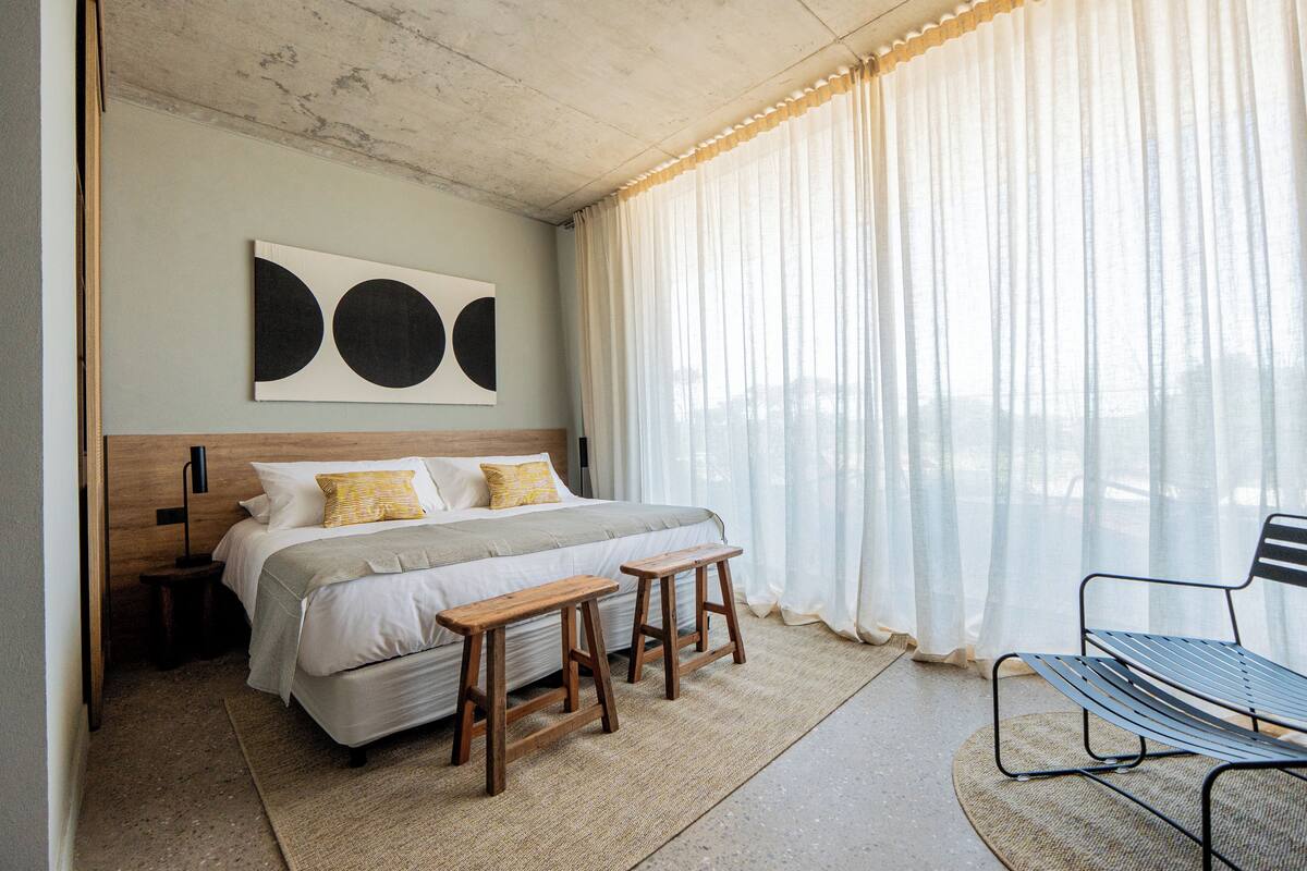A bedroom with a bed, wooden stools, a bench, a wall-mounted artwork, and sheer curtains.