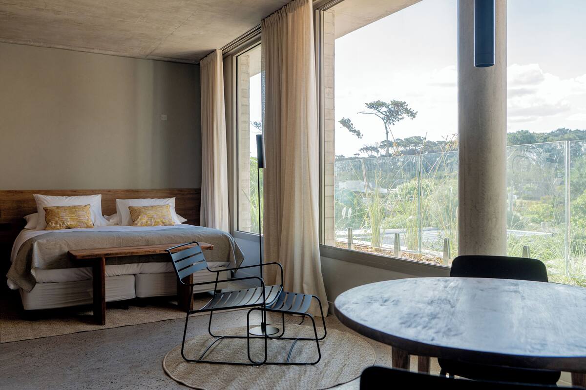 A hotel room with a bed, a wooden bench, a round table, and a view of greenery outside.